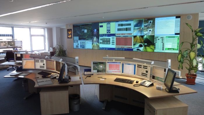 Über eine Wand der Verkehrsrechnerzentrale erstreckt sich eine Vielzahl an Bildschirmen mit unterschiedlichen Anzeigen zur Verkehrssteuerung. Davor befinden sich mehrere Operatoren-Arbeitsplätze.